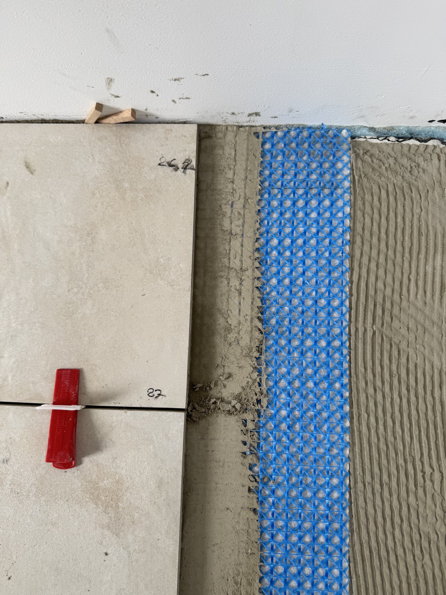 Detail des Bodenaufbaus: verlegte Fliese, aufgekämmtes Kleberbett und darunter die blaue Entkopplungsmatte