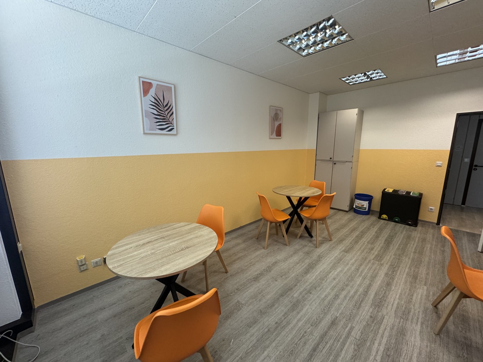 Aufenthaltsraum mit weißer Wandfläche und ockerfarbenem Sockel, runde Tische und orange Stühle.